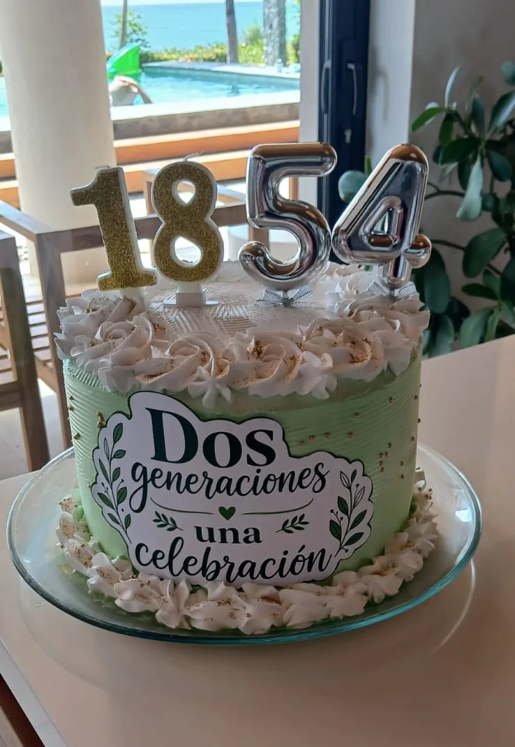 Queque verde con rosetones crema, velas de los números 18 y 54 y un letrero que dice «Dos generaciones, una celebración»